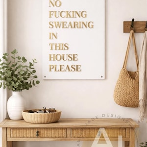 May include: A white framed sign with gold lettering that reads "NO FUCKING SWEARING IN THIS HOUSE PLEASE" hangs on a wall. A woven bag and scarf hang on a wooden rack. A wooden console table holds a vase, basket, and sneakers.