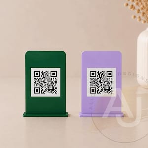 May include: Two small, rectangular QR code stands. One is dark green, the other is lavender. Each stand has a white square with a black QR code. The stands are on a light-coloured surface.