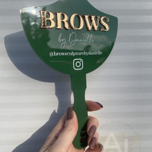Op de afbeelding: Een groene handspiegel met een gouden inscriptie die "Brows by Danielle" luidt en de social media-handle "@browsculpturebydanielle". De spiegel is waaiervormig en heeft een groene handgreep.