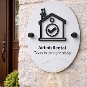 May include: White oval Airbnb Rental sign with black detailing, fixed to a stone wall. The sign shows a house icon with a tick, the text "Airbnb Rental, You're in the right place!" and the "JADE DESIGN" logo.