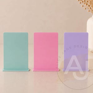 May include: Three rectangular display stands in pastel colors: mint green, pink, and lavender. The stands have a base and a vertical panel. The lavender stand has the text "ALICE JADE DESIGNS" and the letters "AJ" printed on it.