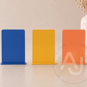Op de afbeelding: Drie kleurrijke acrylborden in blauw, geel en oranje. Elk bord heeft een rechthoekige vorm met afgeronde hoeken en een voet. Het oranje bord heeft de tekst "ALICE JADE DESIGNS" en de letters "AJ".