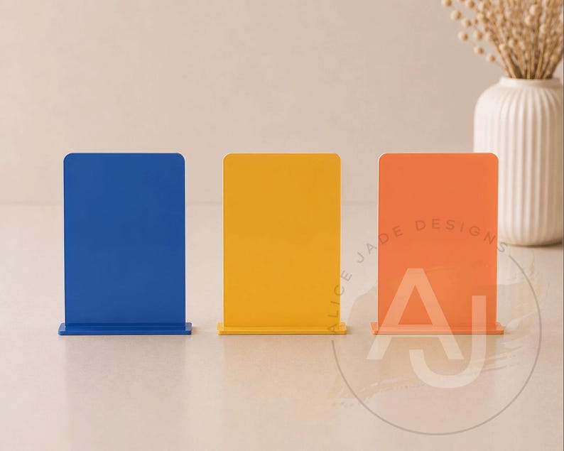 Op de afbeelding: Drie kleurrijke acrylborden in blauw, geel en oranje. Elk bord heeft een rechthoekige vorm met afgeronde hoeken en een voet. Het oranje bord heeft de tekst "ALICE JADE DESIGNS" en de letters "AJ".