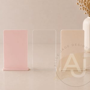 Op de afbeelding: Drie acryl tafelborden in roze, transparant en beige. Het beige bord heeft de tekst "ALICE JADE DESIGNS" en de letters "AJ" in een cirkel. Een witte vaas met gedroogde bloemen staat op de achtergrond.
