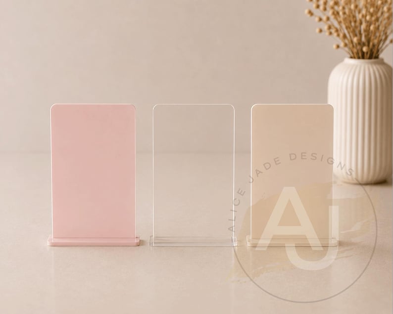 Op de afbeelding: Drie acryl tafelborden in roze, transparant en beige. Het beige bord heeft de tekst "ALICE JADE DESIGNS" en de letters "AJ" in een cirkel. Een witte vaas met gedroogde bloemen staat op de achtergrond.