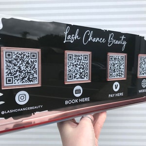 Peut inclure: Panneau en acrylique transparent avec fond noir et bordure or rose. Le panneau affiche le texte "Lash Chance Beauty" et quatre codes QR, avec des icônes pour Instagram, la réservation, le paiement et le site Web.