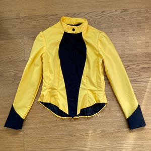 May include: A yellow long-sleeved shirt with a black collar and black trim on the cuffs and down the front. The shirt has a fitted silhouette.