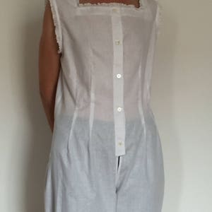 May include: A white sleeveless button-up top with a square neckline and a frilled trim. The top has a long, flowing silhouette and is made of a lightweight fabric.