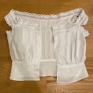 May include: A white, open-front blouse with intricate lace detailing along the neckline and sleeves. The blouse features vertical pleats and delicate ribbon ties, creating a vintage or Victorian aesthetic. The sleeves are short and gathered at the cuffs.