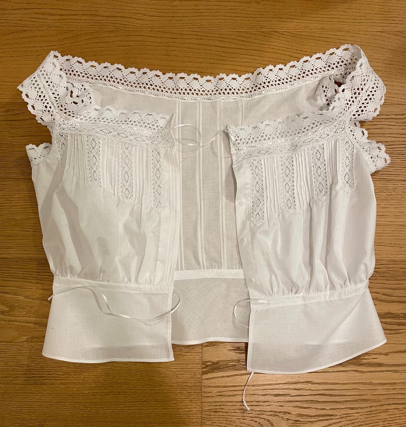 May include: A white, open-front blouse with intricate lace detailing along the neckline and sleeves. The blouse features vertical pleats and delicate ribbon ties, creating a vintage or Victorian aesthetic. The sleeves are short and gathered at the cuffs.