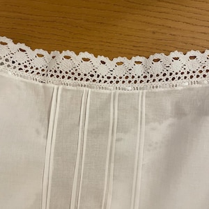 May include: Close-up of a white garment with a decorative, scalloped lace trim along the neckline. Vertical pleats and subtle stripes run down the front of the fabric, creating a textured effect. The background is a warm, wooden surface.
