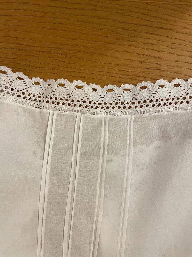 May include: Close-up of a white garment with a decorative, scalloped lace trim along the neckline. Vertical pleats and subtle stripes run down the front of the fabric, creating a textured effect. The background is a warm, wooden surface.