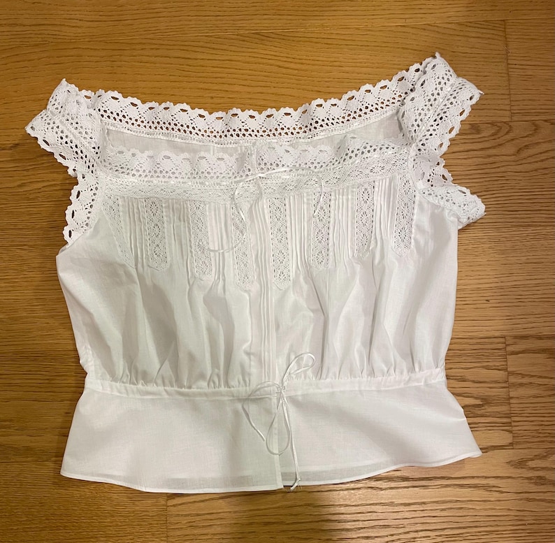May include: White cotton top with lace trim and detailing. The top features a square neckline, short sleeves, and a drawstring waist. The bodice has vertical pleats and lace inserts, creating a delicate and feminine look. The top is laid flat on a wooden surface.