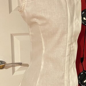 May include: A white linen top with a lace trim around the neckline and armholes. The top is fitted and has a slightly gathered waist.