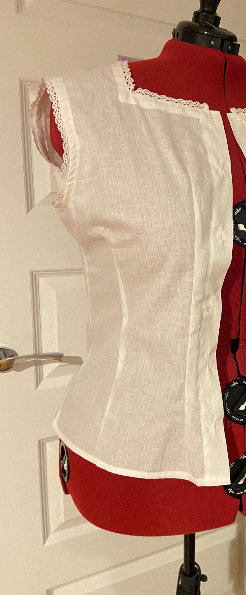 May include: A white linen top with a lace trim around the neckline and armholes. The top is fitted and has a slightly gathered waist.