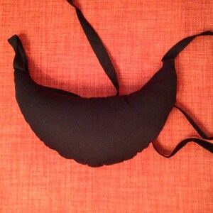 May include: A black crescent-shaped pillow with a long black strap. The pillow is lying on a red textured surface.