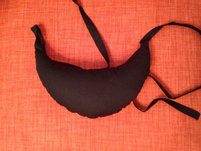 May include: A black crescent-shaped pillow with a long black strap. The pillow is lying on a red textured surface.