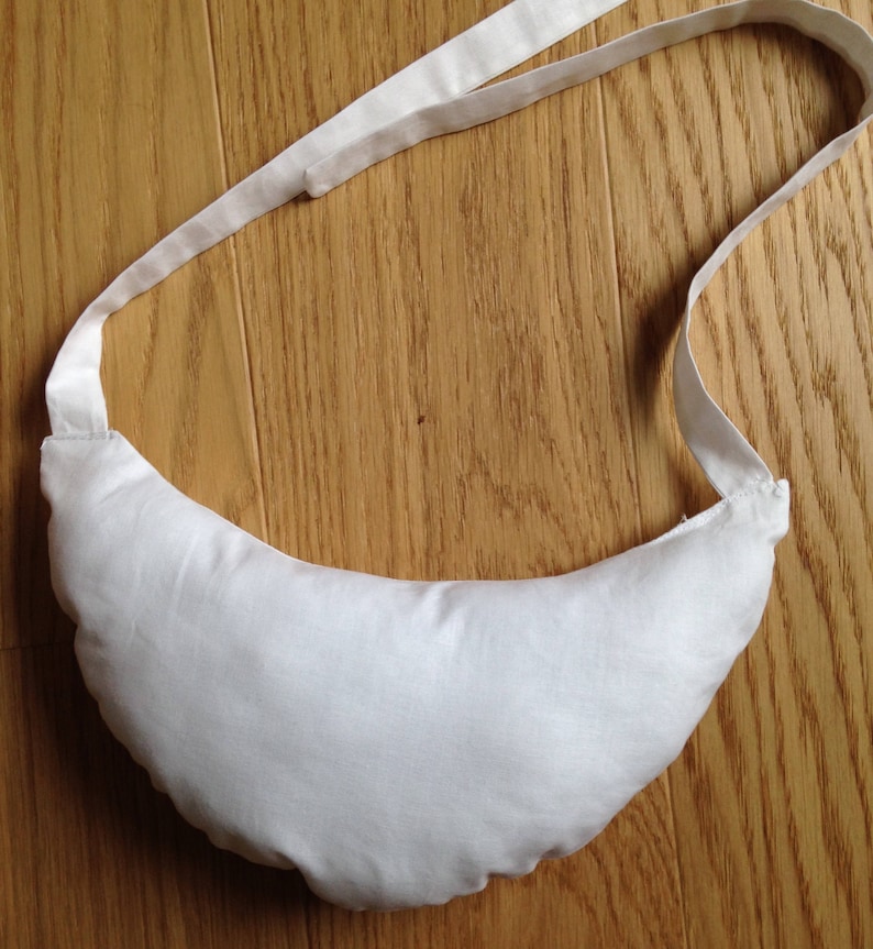 May include: A white crescent-shaped pillow with a white fabric strap. The pillow is made of soft, white fabric and has a simple design.