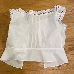 May include: A white sleeveless top with a delicate lace trim along the neckline and shoulders. The bodice features vertical pleats, and the bottom has a flared, ruffled hem. The fabric appears lightweight and semi-sheer, suggesting a vintage or romantic style.