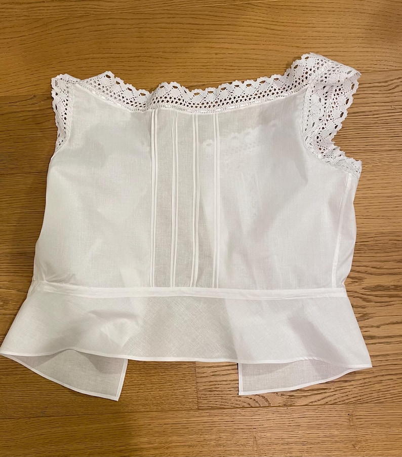 May include: A white sleeveless top with a delicate lace trim along the neckline and shoulders. The bodice features vertical pleats, and the bottom has a flared, ruffled hem. The fabric appears lightweight and semi-sheer, suggesting a vintage or romantic style.