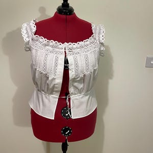 May include: White, off-the-shoulder blouse with lace trim and detailing. The blouse is displayed on a red dress form. The top features a square neckline and button-up front. The sleeves are short and trimmed with lace.