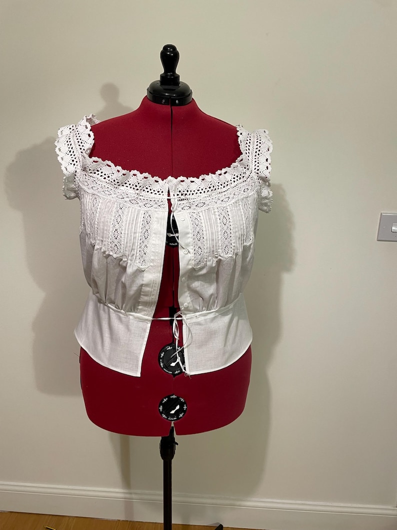 May include: White, off-the-shoulder blouse with lace trim and detailing. The blouse is displayed on a red dress form. The top features a square neckline and button-up front. The sleeves are short and trimmed with lace.