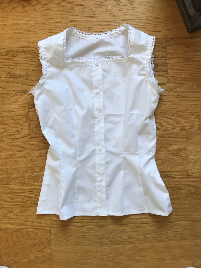 May include: A white sleeveless button-up blouse with a square neckline and lace trim.