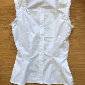 May include: A white sleeveless blouse with a square neckline and a button-up front. The neckline and armholes are trimmed with white lace.