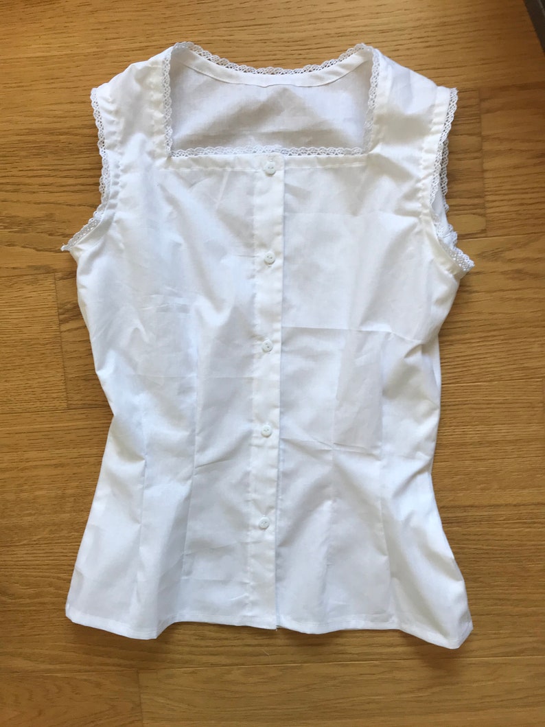 May include: A white sleeveless blouse with a square neckline and a button-up front. The neckline and armholes are trimmed with white lace.