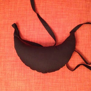 May include: A black crescent-shaped pillow with a black fabric tie. The pillow is on a red textured surface.