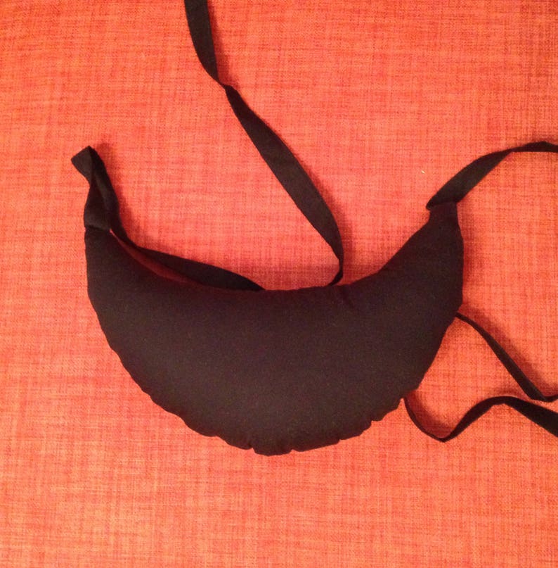 May include: A black crescent-shaped pillow with a black fabric tie. The pillow is on a red textured surface.