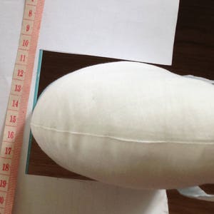May include: A white fabric cushion with a slight curve, being held by a hand. The cushion is being placed on a white surface with a ruler marked in inches along the left side.