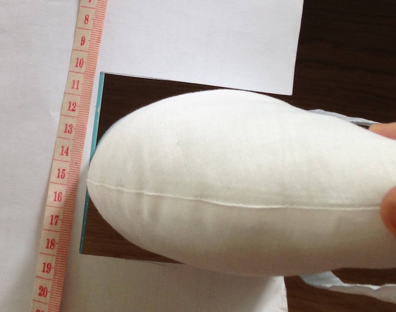May include: A white fabric cushion with a slight curve, being held by a hand. The cushion is being placed on a white surface with a ruler marked in inches along the left side.