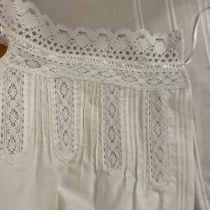 May include: White cotton top with crochet lace trim on the neckline and vertical panels. The top has tiny pleats.