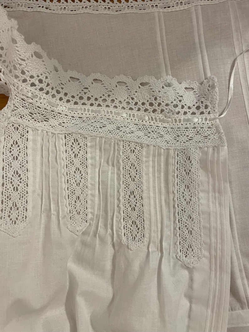 May include: White cotton top with crochet lace trim on the neckline and vertical panels. The top has tiny pleats.