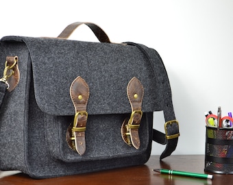 May include: A gray felt messenger bag with brown leather accents and brass buckles. The bag has a top handle and an adjustable shoulder strap.