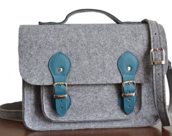May include: A gray felt satchel bag with teal leather accents and buckles. The bag has a top handle and a long adjustable shoulder strap.