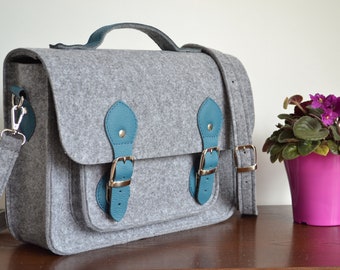 May include: A gray felt satchel bag with teal leather accents and buckles. The bag has a long shoulder strap and a top handle.