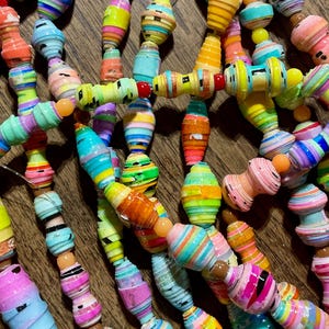 May include: Close-up of colorful paper bead necklaces. The beads are cylindrical and barrel-shaped, with vibrant stripes of pink, yellow, blue, and orange. The necklaces are strung together with a thin, clear cord.