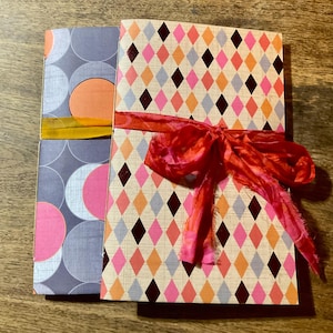 May include: Two decorative journals with patterned covers. One journal has a gray background with pink and orange circles. The other journal has a diamond pattern in pink, orange, and black, tied with a red ribbon.
