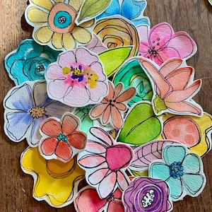May include: A collection of colorful, hand-painted watercolor flower cutouts. The flowers feature a variety of shapes and colors, including yellow, pink, blue, and orange, with black outlines. Green leaves are also included in the assortment.