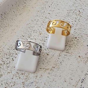 Può includere: Due anelli, uno dorato e uno argentato, esposti su supporti bianchi. Entrambi gli anelli presentano un design continuo a forma di onda. L'anello dorato ha una finitura lucida, mentre l'anello argentato ha un aspetto lucido.