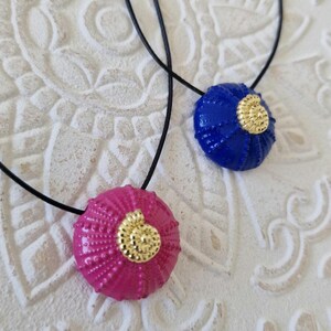 Könnte beinhalten: Zwei Anhänger-Halsketten mit runden, strukturierten Anhängern. Ein Anhänger ist hellrosa, der andere blau. Jeder hat ein goldfarbenes Spiraldesign in der Mitte und hängt an einer schwarzen Kordel. Der Hintergrund ist eine weiße, gemusterte Oberfläche.