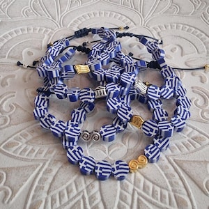 Op de afbeelding: Verschillende armbanden met blauw-witte kralen met het thema van de Griekse vlag. De armbanden hebben verstelbare zwarte koorden en goud- of zilverkleurige accenten. De kralen zijn zeshoekig.