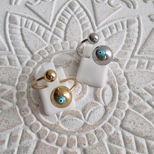 May include: Two rings with a unique design. One ring is gold-toned with a large, round charm featuring an eye design with a blue center. The other ring is silver-toned with a similar eye charm. Both rings have a small ball on top.
