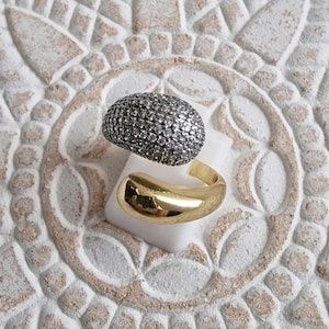 May include: A bypass ring featuring two distinct ends. One end is a rounded, dome-shaped element covered in numerous small, clear, sparkling stones. The other end is a smooth, polished, solid gold-toned metal curve that tapers to a point.