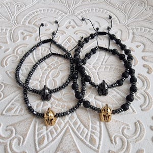 May include: Four adjustable bracelets featuring spherical beads. Two bracelets consist of metallic gray hematite beads with a central gold or black Spartan helmet charm. Two bracelets feature black porous lava beads and metallic gray spacers.