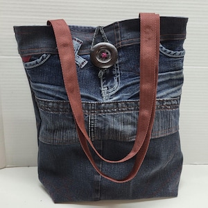Peut inclure: Un sac cabas en jean bleu avec des anses marron et une grande fermeture à bouton. Le sac est fabriqué à partir de denim recyclé et présente un design patchwork.