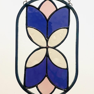 May include: Stained glass window panel with a floral design. The panel features blue, pink, and off-white glass pieces set within a black metal frame. A chain is attached for hanging. The design is symmetrical and abstract.