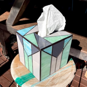 May include: A geometric stained glass tissue box in shades of blue, green, grey and white. The box has dark grey outlines and contains white tissues. The box is sitting on a wooden stool.